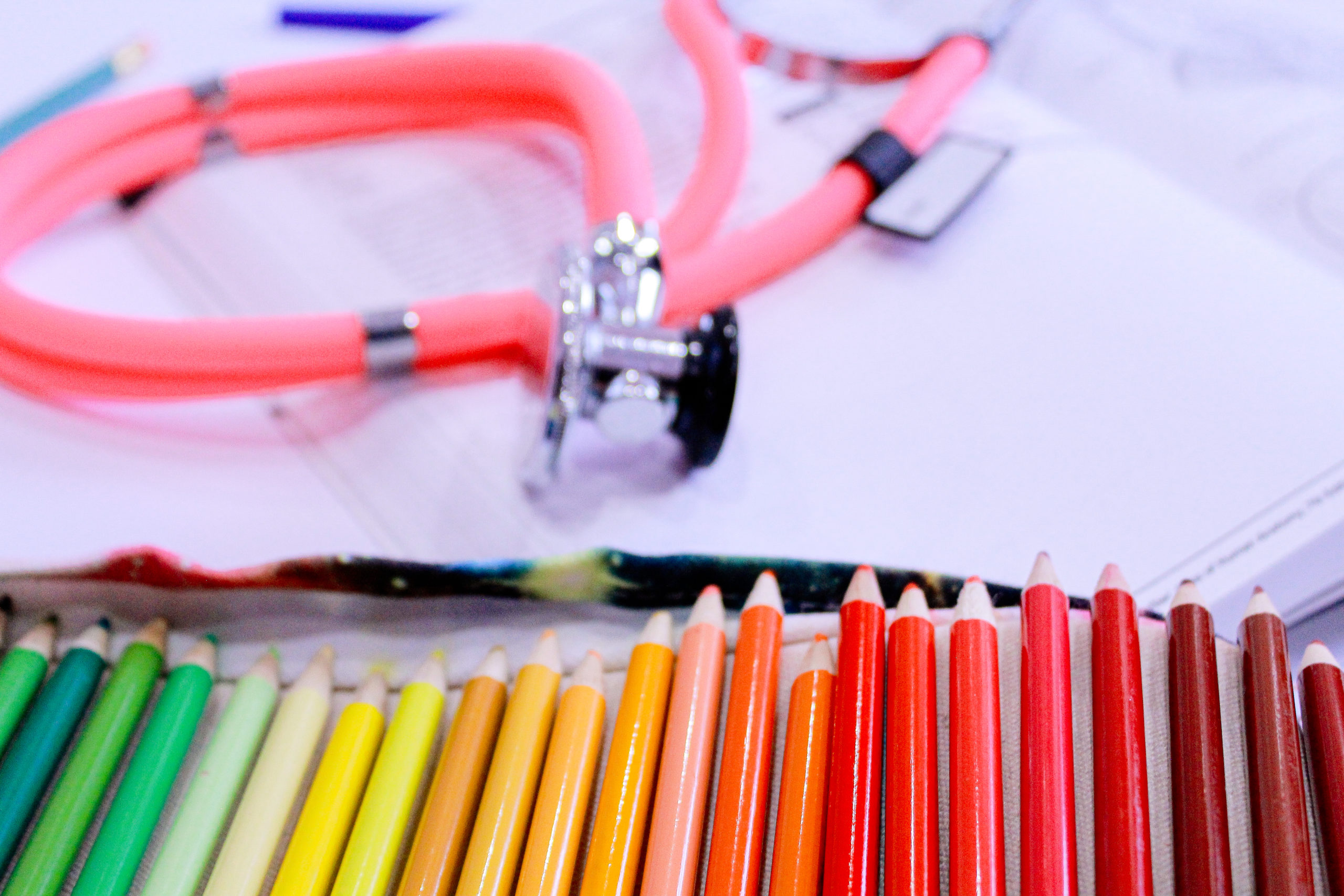 "pink stethoscope on top of Netter Anatomy Coloring Book next to roll of colored pencils in rainbow order, green colored pencils on left to red on right"